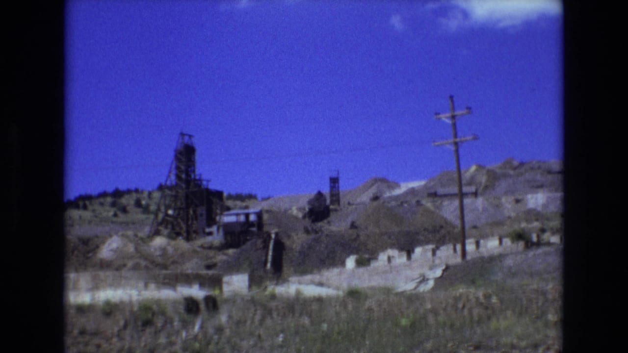 1973 Colorado Springs-area 8mm frame showing a dark mine shaft structure amid excavated rock, a wooden utility pole, and mountainous terrain under a strong blue sky.