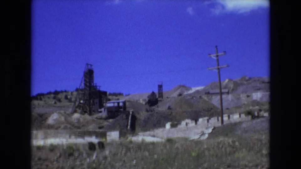 1973 Colorado Springs-area 8mm frame showing a dark mine shaft structure amid excavated rock, a wooden utility pole, and mountainous terrain under a strong blue sky.