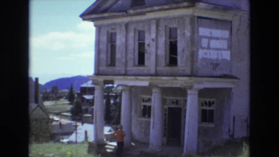1973 Colorado Springs-area 8mm frame showing an old two-story columned building with open windows, hillside scenery, and a small person standing near the front steps.