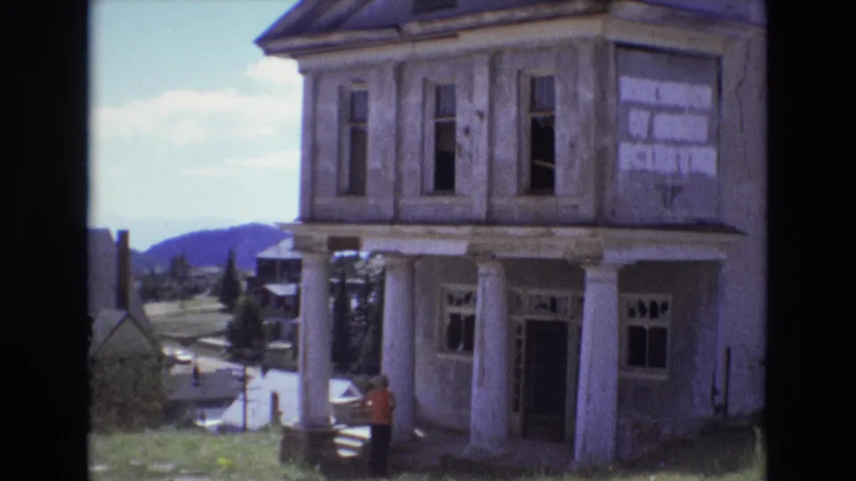 1973 Colorado Springs-area home-movie frame of an old columned building with empty window openings, grass in front, distant hills, and a small visitor figure at the steps.