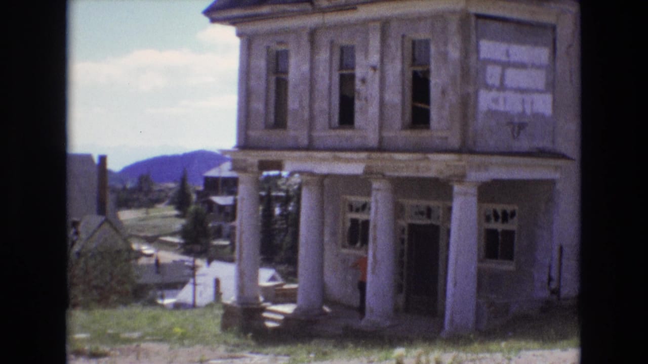1973 Colorado Springs home-movie frame showing a weathered two-story columned building with broken windows, grass in front, distant hills, and a small human figure near the entrance.