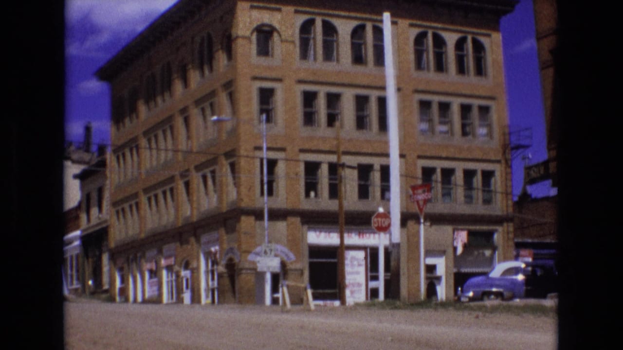 Brick corner commercial building with storefront signs, stop signs, and a parked vintage car.