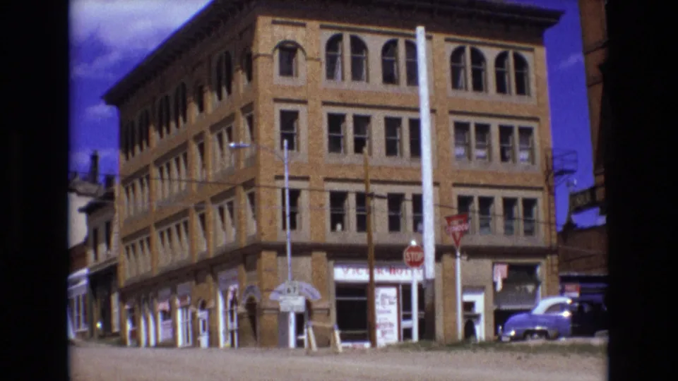 1973 Colorado Springs home-movie frame with a brick corner building filling the view, showing arched upper windows, lower storefronts, street signs, poles, and a parked car at the curb.