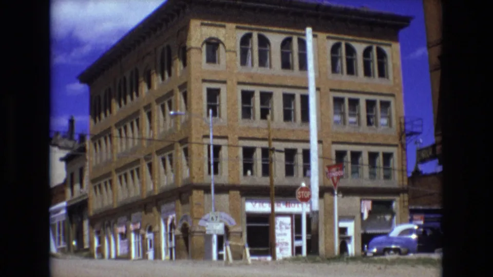 1973 Colorado Springs frame showing the same multi-story brick commercial building from a slightly shifted angle, with arched windows, visible shop signs, stop signs, utility poles, and a purple vintage car.