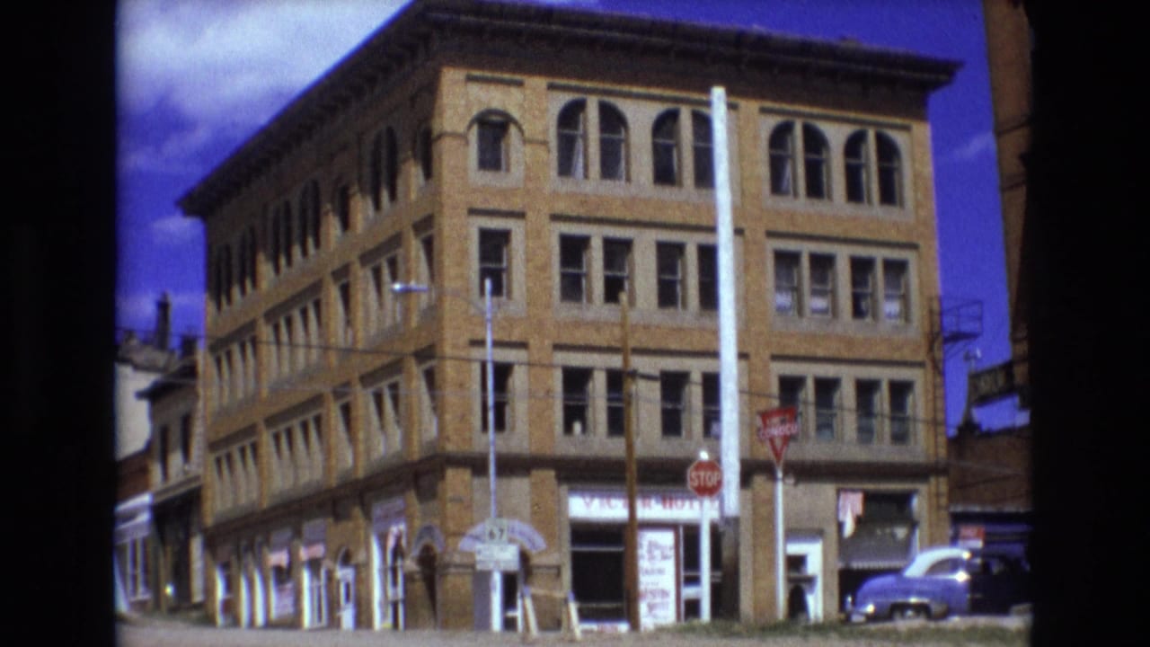 1973 Colorado Springs street view with a multi-story brick corner building, arched windows, stop signs, storefronts, and a car near the curb.