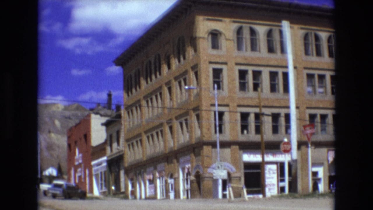 1973 Colorado Springs town street showing a brick commercial building, stop signs, older cars, and low mountains visible beyond nearby storefronts.