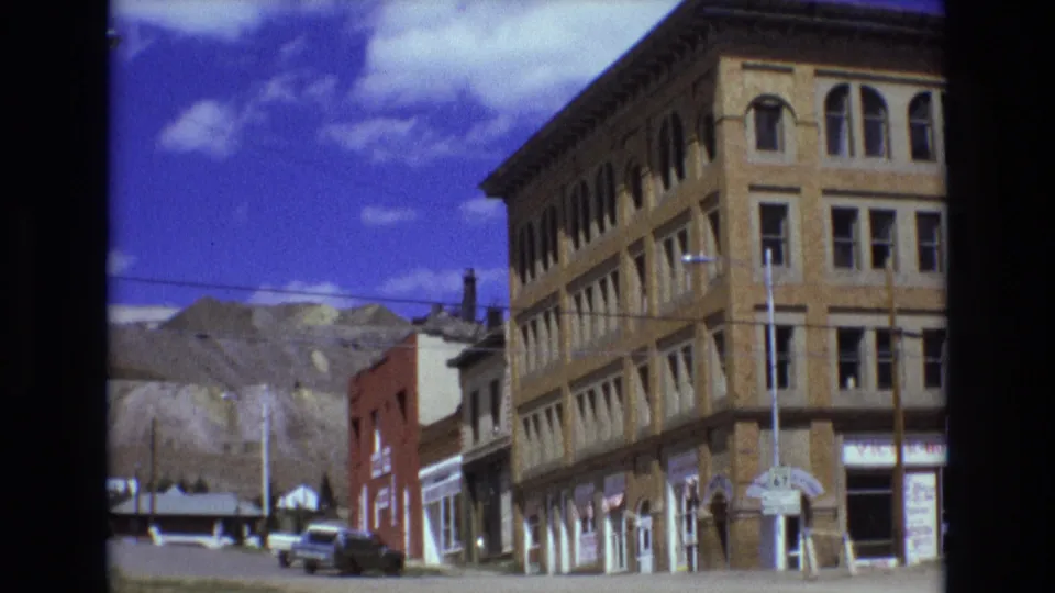 1973 Colorado Springs street view with the brick corner building partly cropped, a red storefront block, older cars, utility lines, and mountains in the distance.