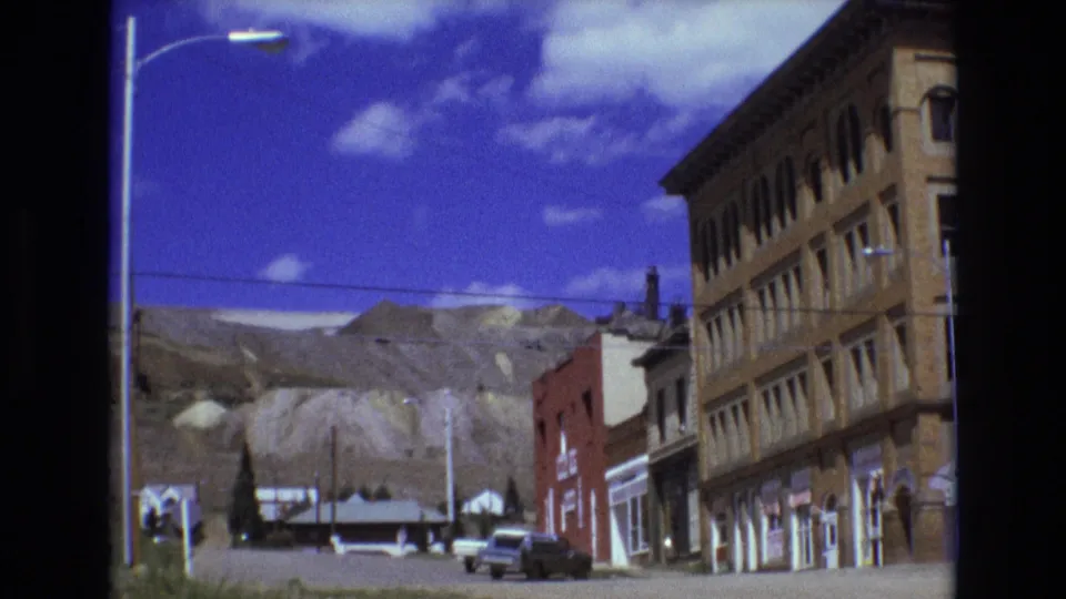 1973 Colorado Springs street scene with mountains behind a low commercial block, older cars near the curb, and the taller brick building partly visible at right.