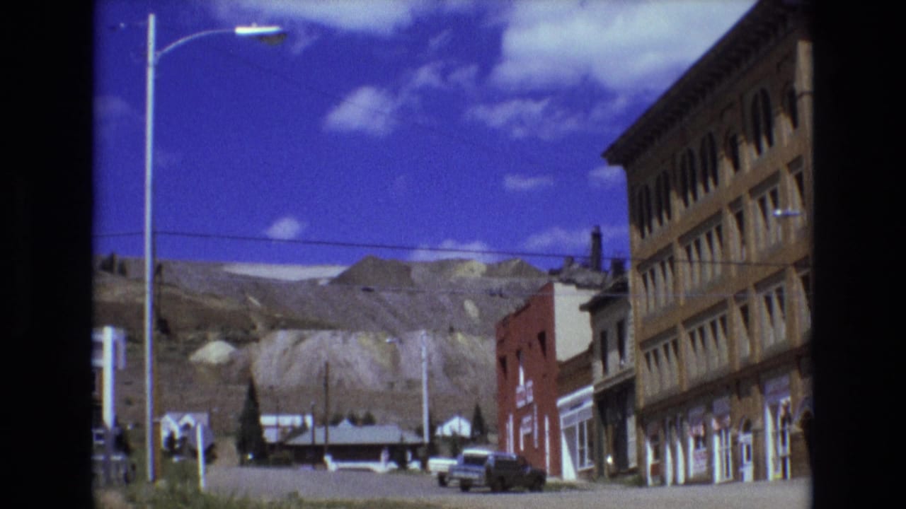 1973 Colorado Springs street view with older commercial buildings, tall streetlights, parked cars, and a rugged mountain backdrop under a blue sky.