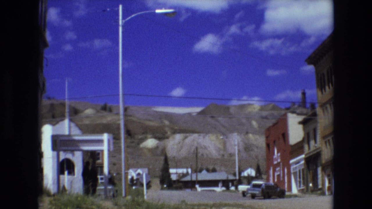 1973 Colorado Springs street scene showing low-rise storefronts, a red brick building, parked cars, tall streetlights, and pale hills behind the town.