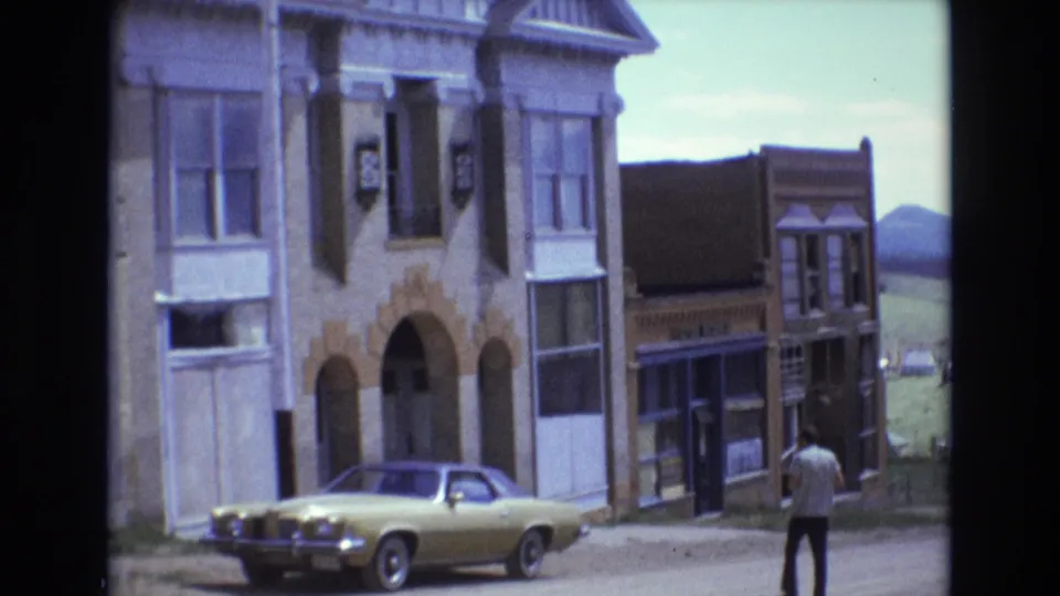 1973 Colorado Springs view of an older stone facade with arched openings, a yellow parked coupe, adjacent storefronts, and a man walking along the sidewalk.