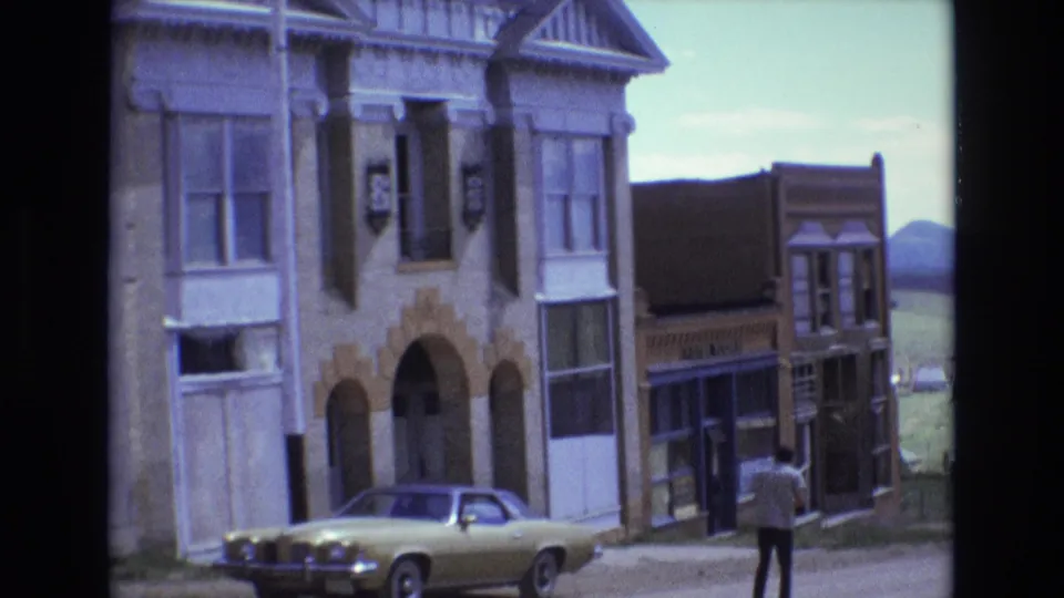 1973 Colorado Springs sidewalk scene with a man walking away from the camera, a yellow parked coupe, older commercial facades, and hills visible beyond the street.