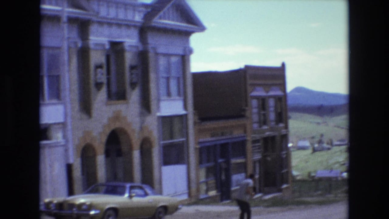 1973 Colorado Springs home-movie frame showing old-style storefront facades, arched windows, a beige sedan, and open hills beyond the buildings.
