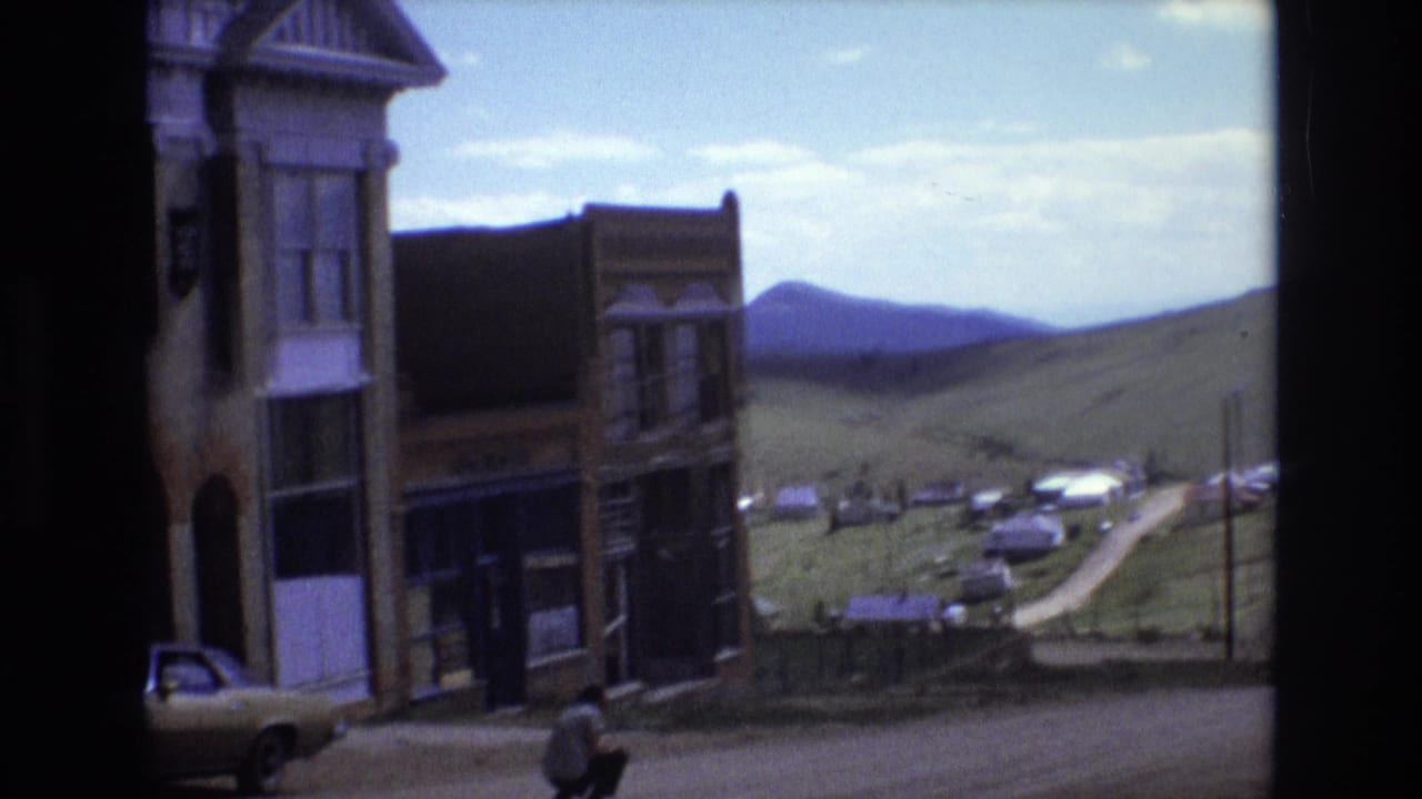 Storefronts, a parked car, a dirt road, and a man crouching near the street.