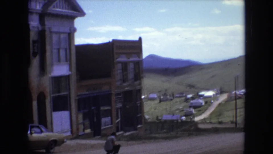 1973 Colorado Springs home-movie view of old commercial facades, a beige car, a crouching adult man, and a road leading toward parked vehicles in the hills.