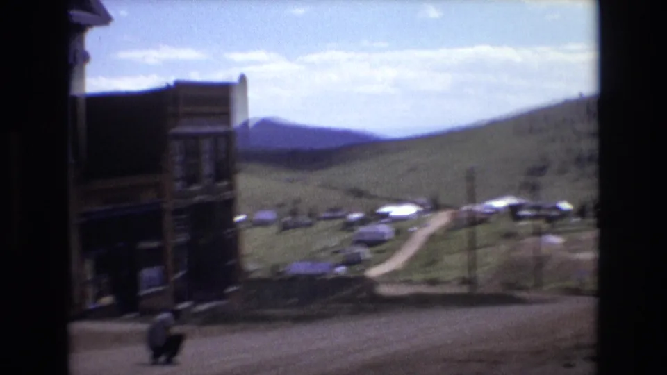1973 Colorado Springs home-movie frame with old storefront edges, a crouching man near the road, distant parked cars, and open green hills under a bright sky.