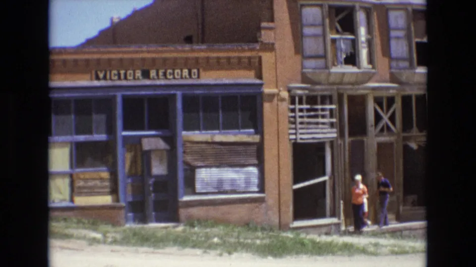 1973 Colorado Springs home-movie frame showing a Victor Record storefront sign, boarded lower windows, upper windows, and two adults standing or walking near the sidewalk.