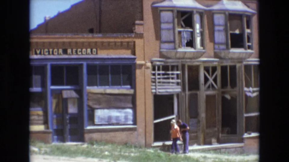 1973 Colorado Springs street scene with two pedestrians in front of the damaged Victor Record storefront, showing boarded sections, broken windows, and bright midday light.