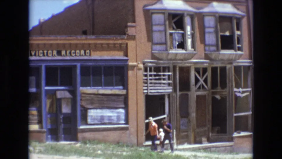 1973 Colorado Springs frame showing two pedestrians near the entrance of a damaged Victor Record storefront with exposed window frames and worn blue-painted trim.