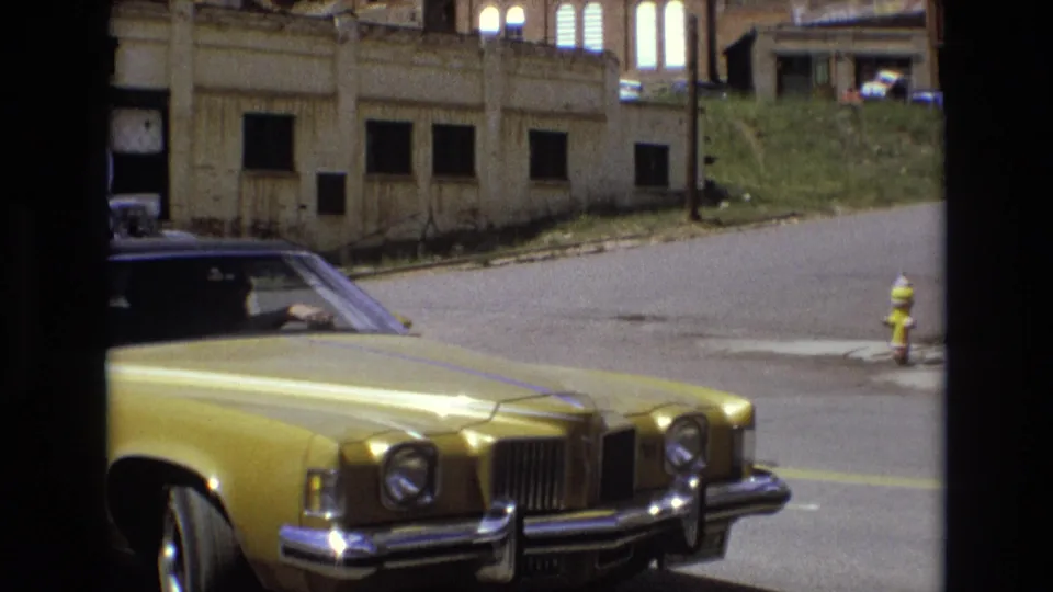 A gold 1970s American sedan is parked along a Colorado Springs street near a fire hydrant, curb, older building facade, and sloped grassy lot.