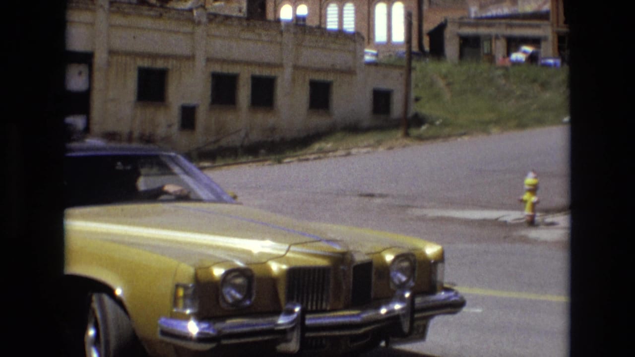 In supported 1973 Colorado Springs home-movie footage, a golden American sedan sits near a curb while worn buildings, a grassy slope, and a yellow fire hydrant appear across the street.