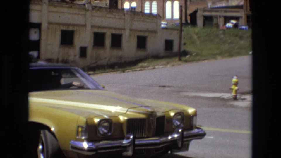 In supported 1973 Colorado Springs home-movie footage, a golden American sedan sits near a curb while worn buildings, a grassy slope, and a yellow fire hydrant appear across the street.