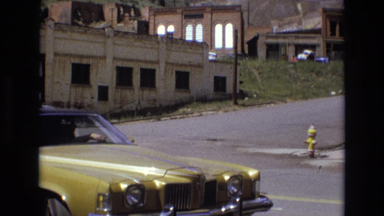 Golden sedan front end beside a street, with boarded buildings and a fire hydrant in the background.