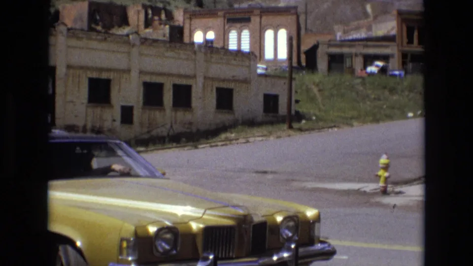 The front of a golden 1970s sedan fills the lower frame while the street, hydrant, grassy bank, and weathered boarded building remain visible behind it.
