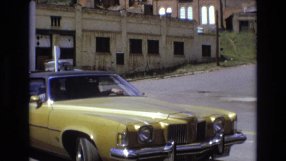 A golden American sedan remains parked in the foreground, seen from behind a dark frame, with boarded openings, masonry walls, and a quiet roadway in the background.