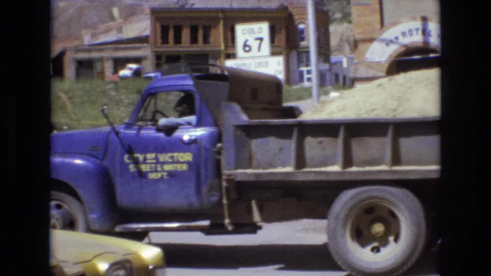 A blue City of Victor Street and Water Department dump truck loaded with gravel passes through the street scene, with the golden car edge, roadside sign, and worn buildings visible nearby.