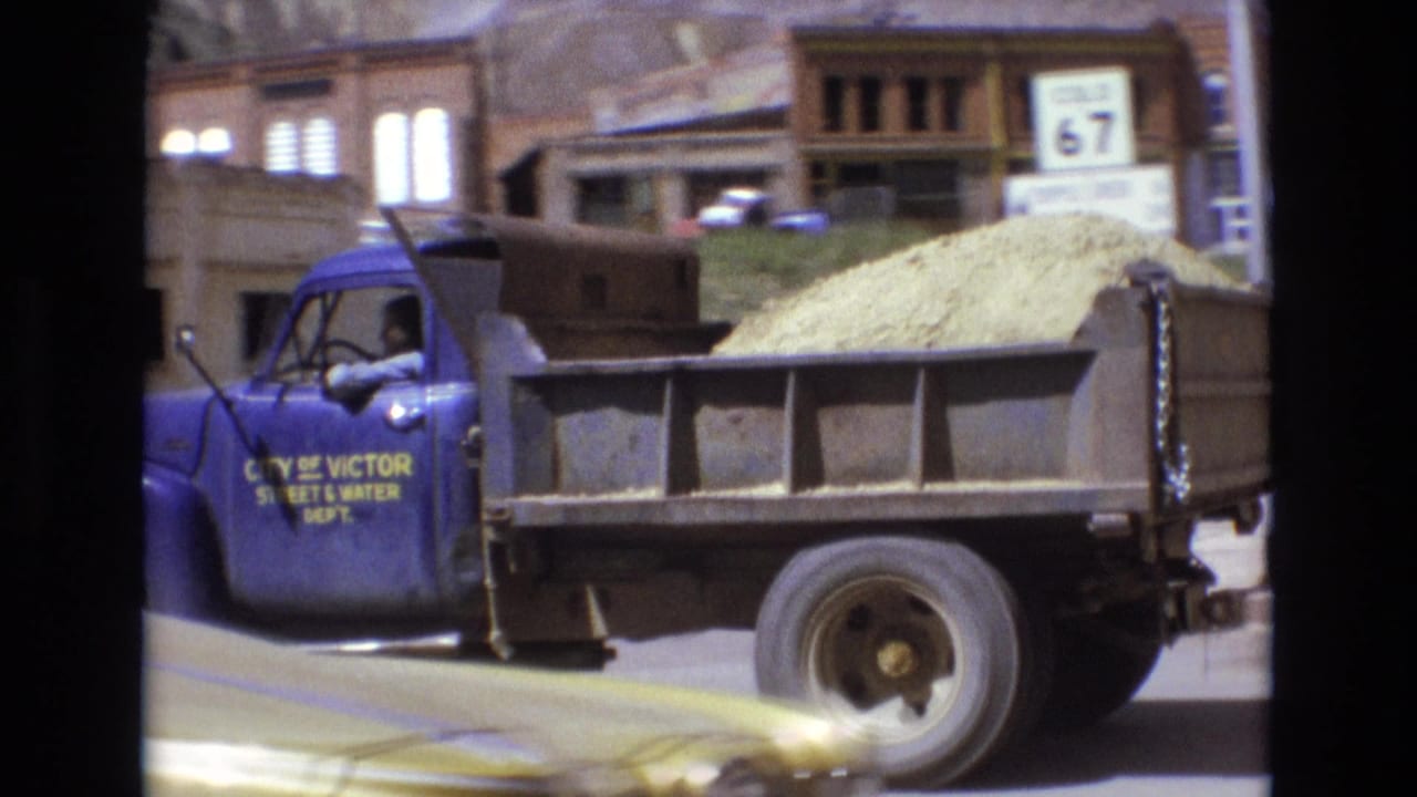 Blue dump truck loaded with sand passes buildings on a sunny street.