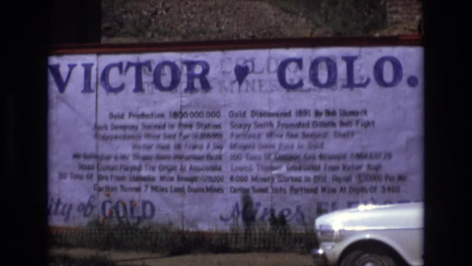 A large Victor, Colo. sign listing gold mining facts remains in view as a white car occupies the lower right corner of the 8mm frame.