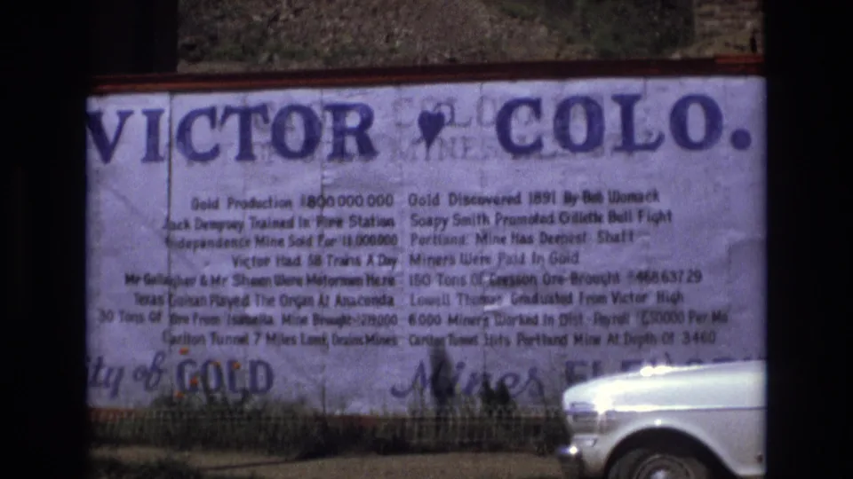 1973 8mm frame of a large Victor, Colorado history sign with mining-related text and a white car partly visible along the bottom edge.