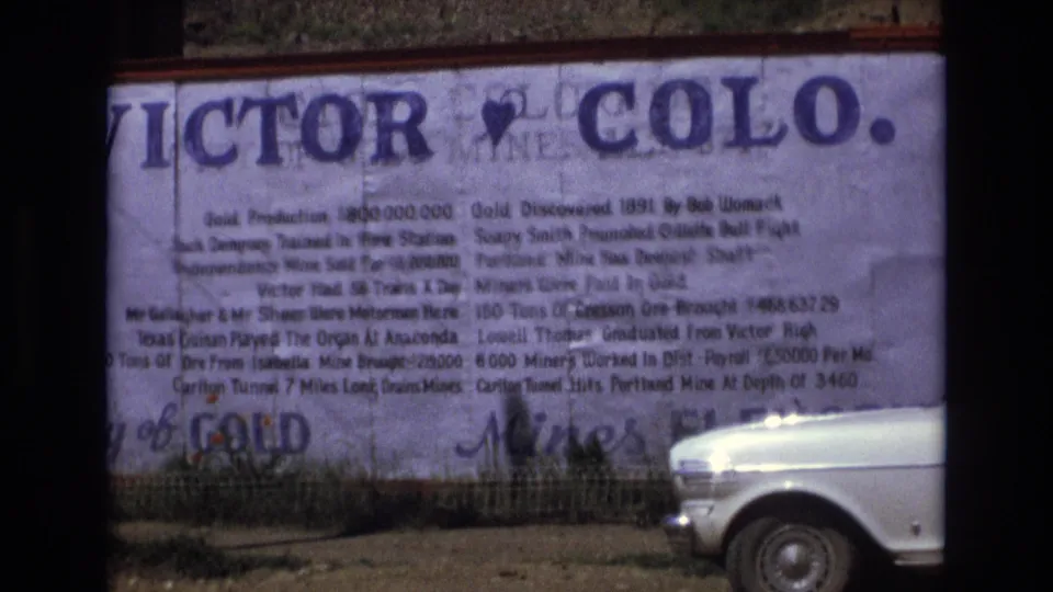 The frame shows the Victor, Colorado sign from a roadside angle, with dense printed text and a white 1970s car at the lower right.
