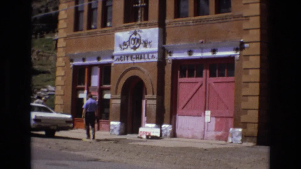 1973 home-movie frame of Victor City Hall, showing its brick facade, red garage-style doors, arched entrance, sidewalk sign, white car, and a man in a blue shirt walking past.