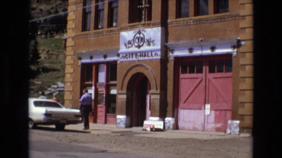 The frame shows Victor City Hall in Victor, Colorado, with red doors, an arched entry, a white sedan, sidewalk signage, and a pedestrian near the doorway.