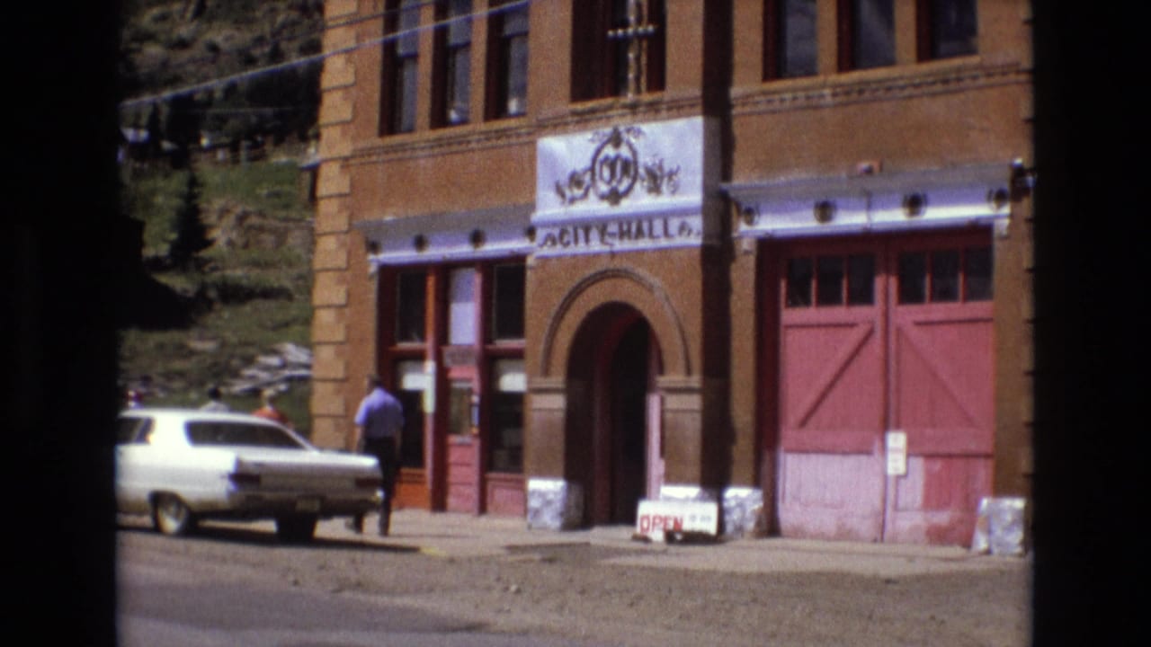 1973 8mm view in Colorado Springs showing a brick City Hall entrance, red doors, a parked white sedan, and a man walking near the sidewalk.