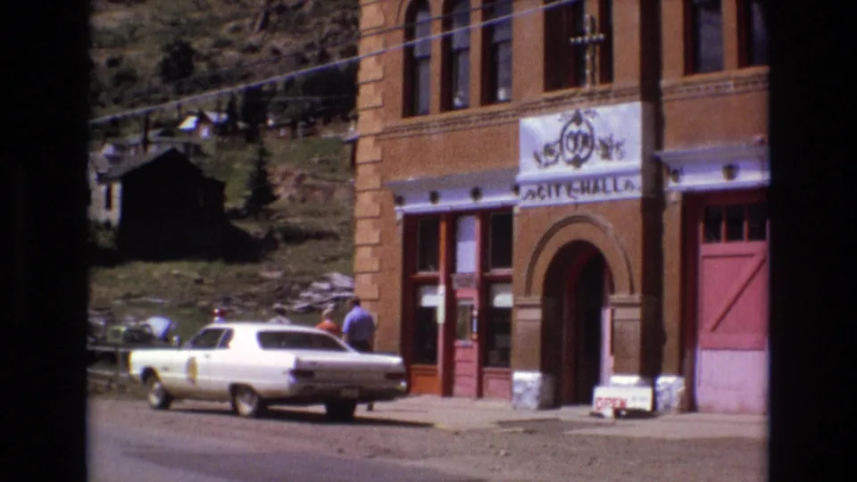 Colorado Springs City Hall exterior with a white 1970s sedan at the curb, a sidewalk sign, red doors, and a man walking in daylight.