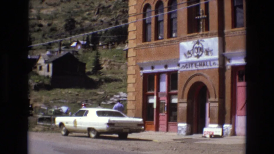 Angled 8mm frame of the Colorado Springs City Hall frontage with the white sedan partly across the lower frame and a man near the doorway.
