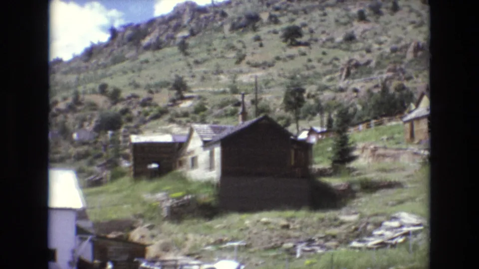 1973 Colorado Springs-area home-movie frame showing small hillside houses, grass, rocks, utility poles, and mountain terrain in soft 8mm focus.