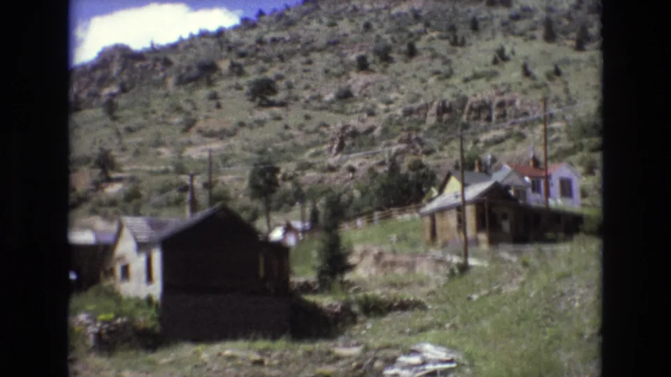 1973 Colorado Springs-area frame of modest hillside homes, utility poles, grass, scattered rocks, and mountain terrain viewed from a handheld home-movie camera.
