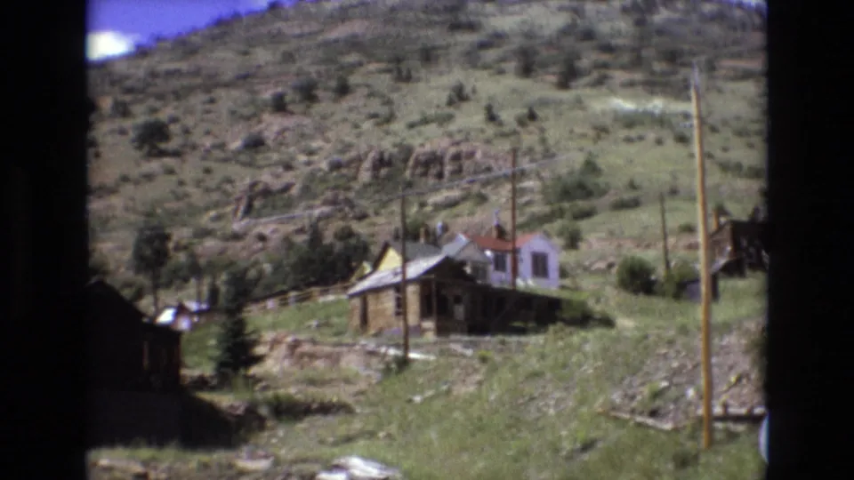 1973 Colorado Springs-area 8mm frame showing a light-colored hillside house with a porch, nearby utility poles, green grass, rocks, and a mountain slope behind it.