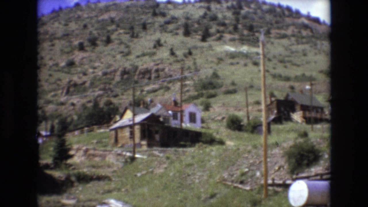 1973 8mm home-movie frame from Colorado Springs showing weathered wooden homes on a grassy hillside beneath a tree-dotted ridge.