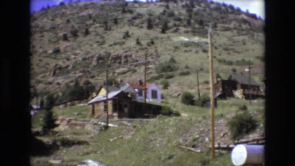 1973 8mm home-movie frame from Colorado Springs showing weathered wooden homes on a grassy hillside beneath a tree-dotted ridge.