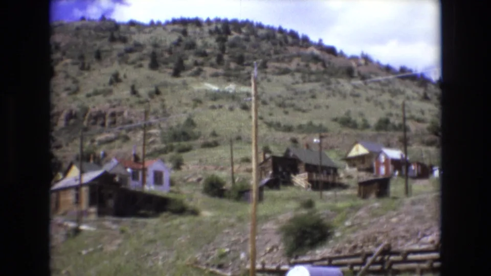 1973 Colorado Springs home-movie frame showing a cluster of rustic wooden buildings, utility poles, and hillside scrub under a partly cloudy sky.