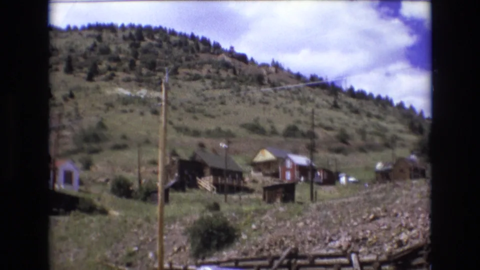 1973 8mm frame from Colorado Springs showing hillside cabins, a rocky foreground, fence rails, utility poles, and scattered trees along the ridge.