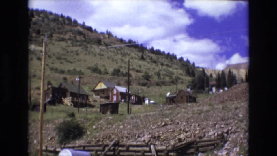 1973 Colorado Springs 8mm frame showing wooden hillside homes, small outbuildings, utility lines, scrub vegetation, and a tree-dotted ridge.