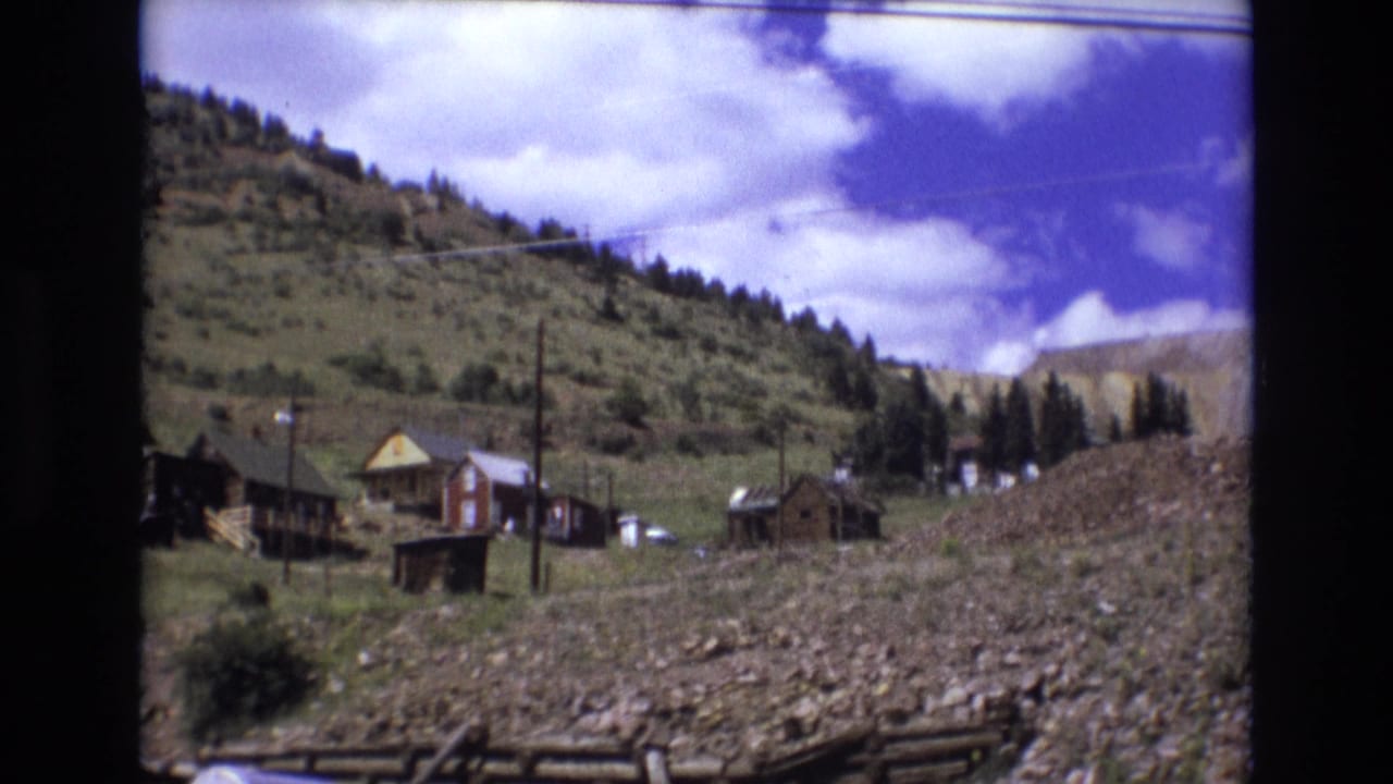 1973 Colorado Springs 8mm frame showing small wooden hillside structures, utility poles, rocky ground, and a pale exposed slope in the background.