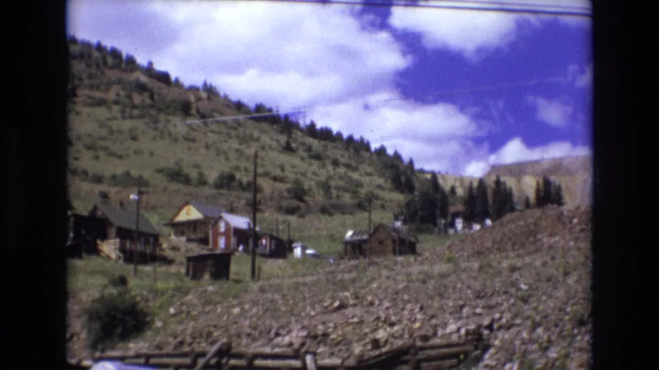 1973 Colorado Springs 8mm frame showing small wooden hillside structures, utility poles, rocky ground, and a pale exposed slope in the background.