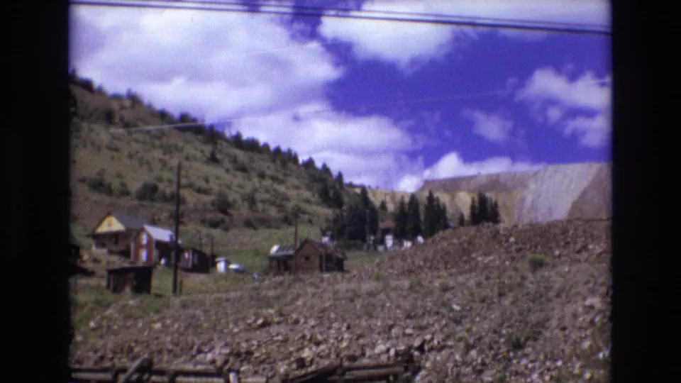 1973 Colorado Springs roadside view of cabins or small structures on a rocky hillside, with trees, utility poles, and a quarry-like face beyond.
